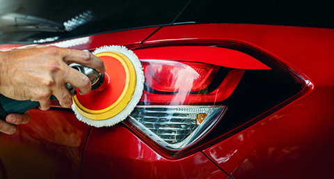 a person buffing the headlight of a red car using a handheld machine polisher